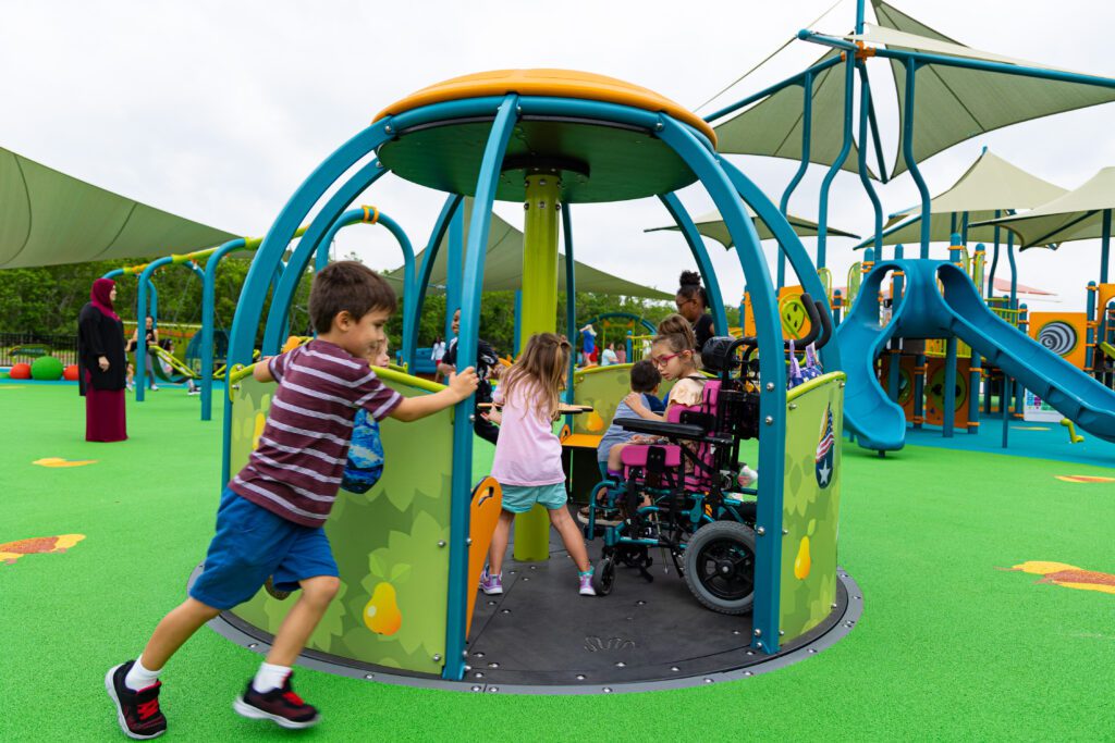 one child pushes an inclusive we-go-round merry go round style spinner at the Ed Thompson Inclusive Park while multiple children spin from within including a child in a wheelchair